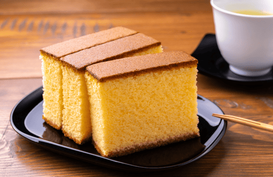 A close-up of a perfectly cut, square slice of Castella, a traditional Japanese sponge cake with a golden-brown top and fluffy yellow interior.