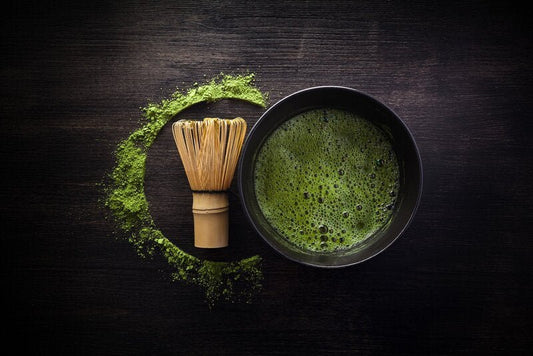 A bamboo whisk next to a bowl of frothy matcha tea with matcha powder spread in a semicircle on a dark wooden surface