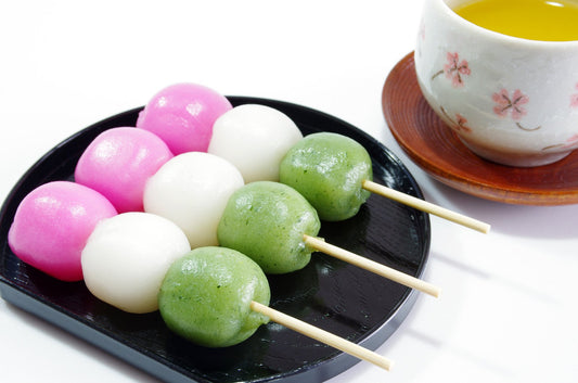 Three-color dango (pink, white, and green) on skewers placed on a black plate. A cup of green tea with a cherry blossom design is in the background.