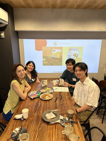 Four people sitting around a wooden table, enjoying snacks and participating in an event, with a presentation screen in the background displaying the JAPANBITE logo.