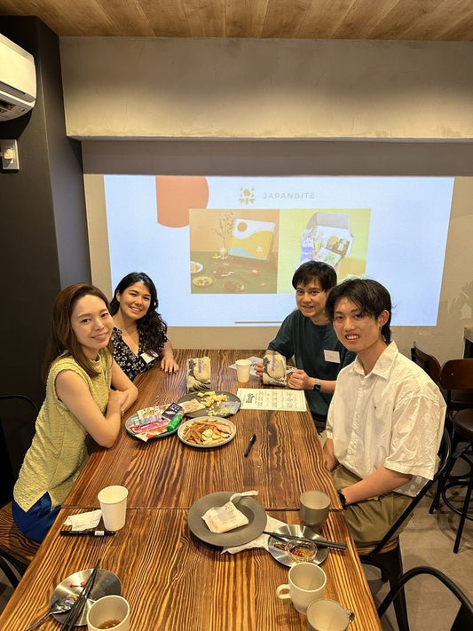 Four people sitting around a wooden table, enjoying snacks and participating in an event, with a presentation screen in the background displaying the JAPANBITE logo.