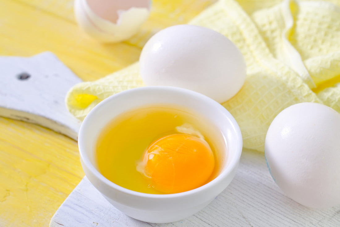 A bowl with a raw egg yolk and white, placed next to whole eggs and a yellow cloth on a light wooden surface.