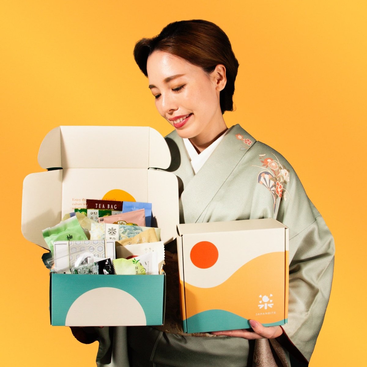 Woman in a traditional Japanese kimono holding open Japanbite gift boxes filled with various snacks and tea, standing against a yellow background.