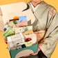 Woman in a traditional Japanese kimono holding an open Japanbite gift box filled with various snacks and tea packages, against a yellow background.